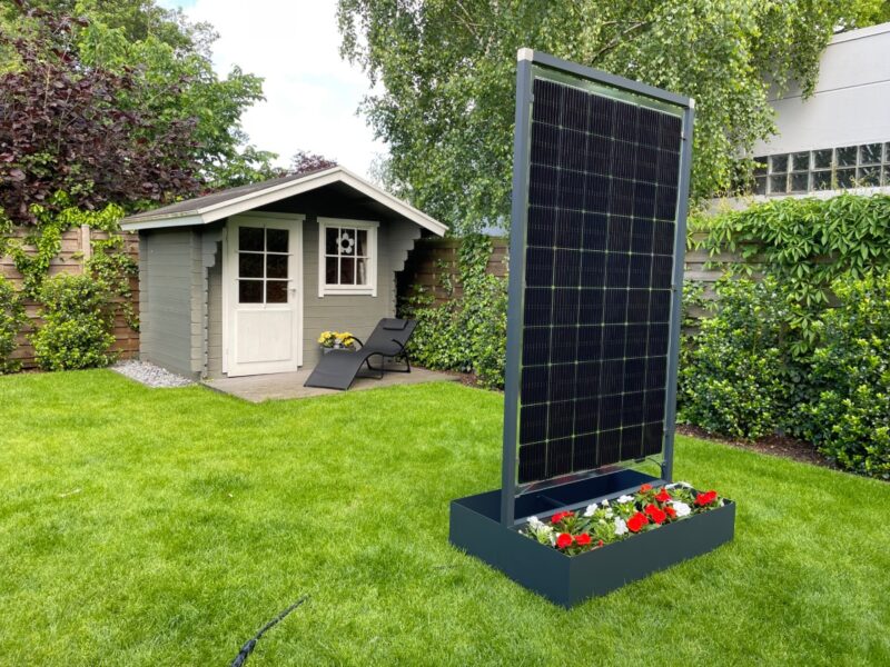 Ein hochkant stehendes Solarmodul mit Blumenkasten vor einem Gartenhäuschen.