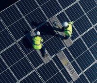 Zwei Menschen in gelben Westen und mit Helm montieren Solarmodule - Symbolbild für Photovoltaik- und Batterieprojekt in Großbritannien.
