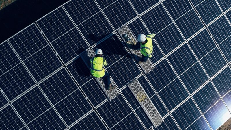 Zwei Menschen in gelben Westen und mit Helm montieren Solarmodule - Symbolbild für Photovoltaik- und Batterieprojekt in Großbritannien.