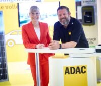 Heike Löffler von Lichtblick und Sascha Coccorullo vom ADAC an einem Stand des ADAC mit PV-Modulen im Hintergrund.