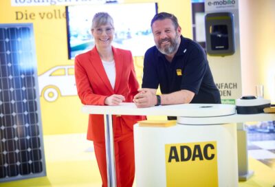 Heike Löffler von Lichtblick und Sascha Coccorullo vom ADAC an einem Stand des ADAC mit PV-Modulen im Hintergrund.