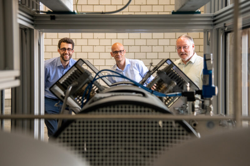 Die drei Verantwortlichen stehen hinter ihrer technischen Anlage und blicken hindurch.
