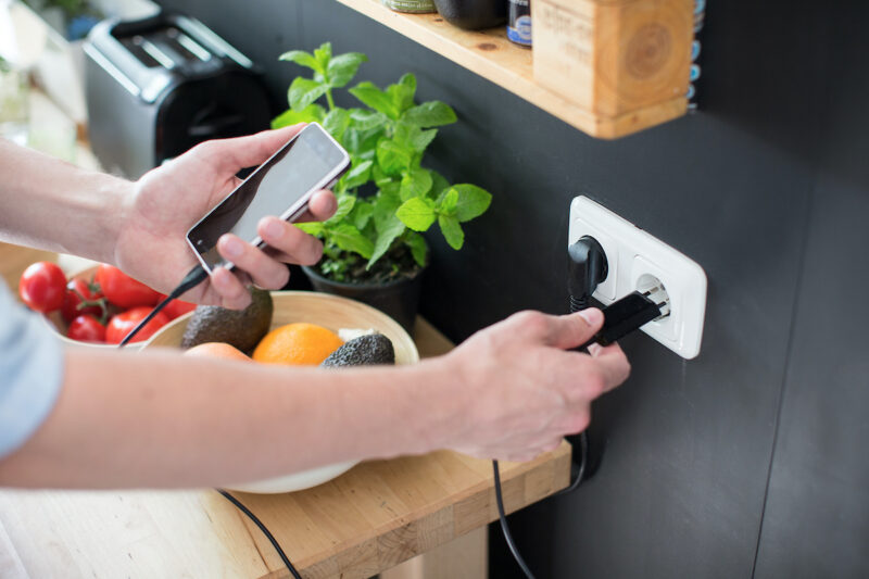 Zu sehen ist in einem Küchenumfeld mit Toaster, Basilikum und Obstkob wie eine linke Hand ein Smartphone hält und eine rechte Hand das Ladekabel in eine weiße Steckdose steckt.