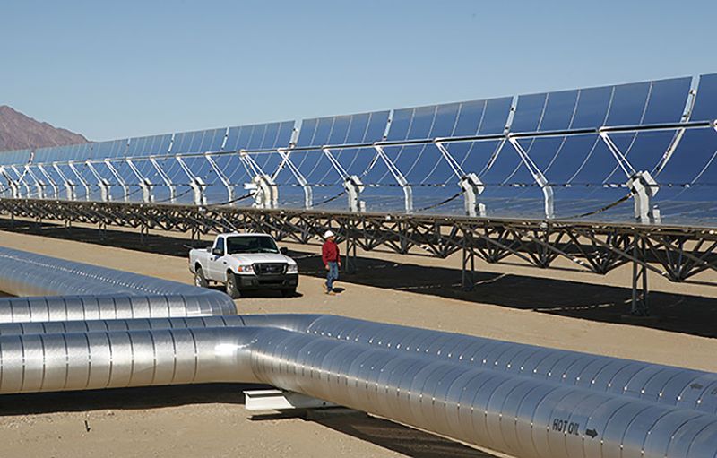 Ein solarthermisches Kraftwerk in der Wüste mit Rohren von Solarspiegeln, dazwischen ein PickUp und Arbeiter mit Helm.