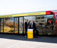 Zwei Männer und ein Ölfass mit Aufschrift Synhelion vor einem Flughafenbus.