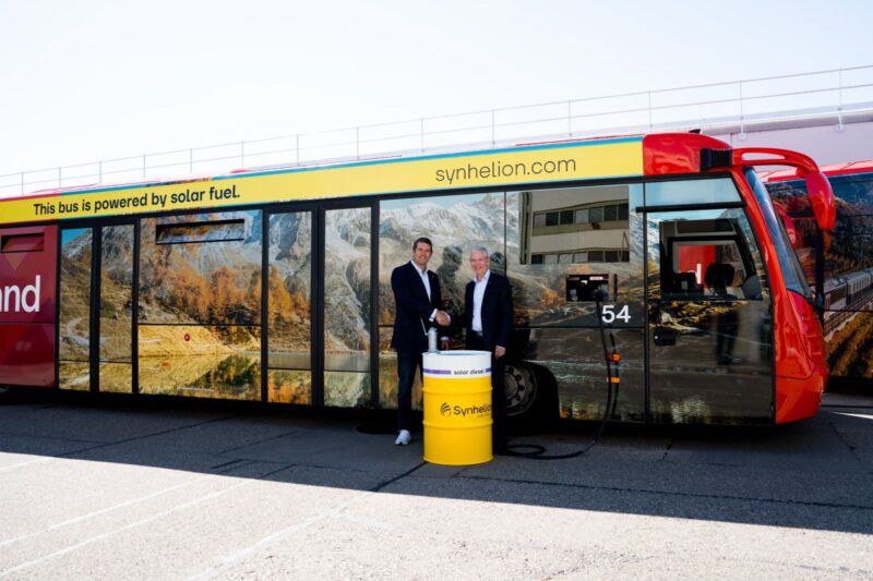 Zwei Männer und ein Ölfass mit Aufschrift Synhelion vor einem Flughafenbus.