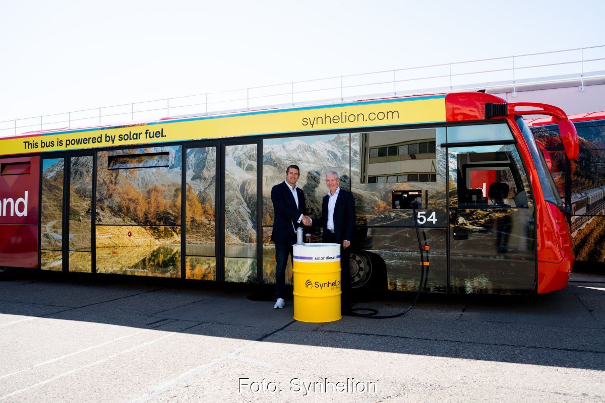Zwei Männer und ein Ölfass mit Aufschrift Synhelion vor einem Flughafenbus.