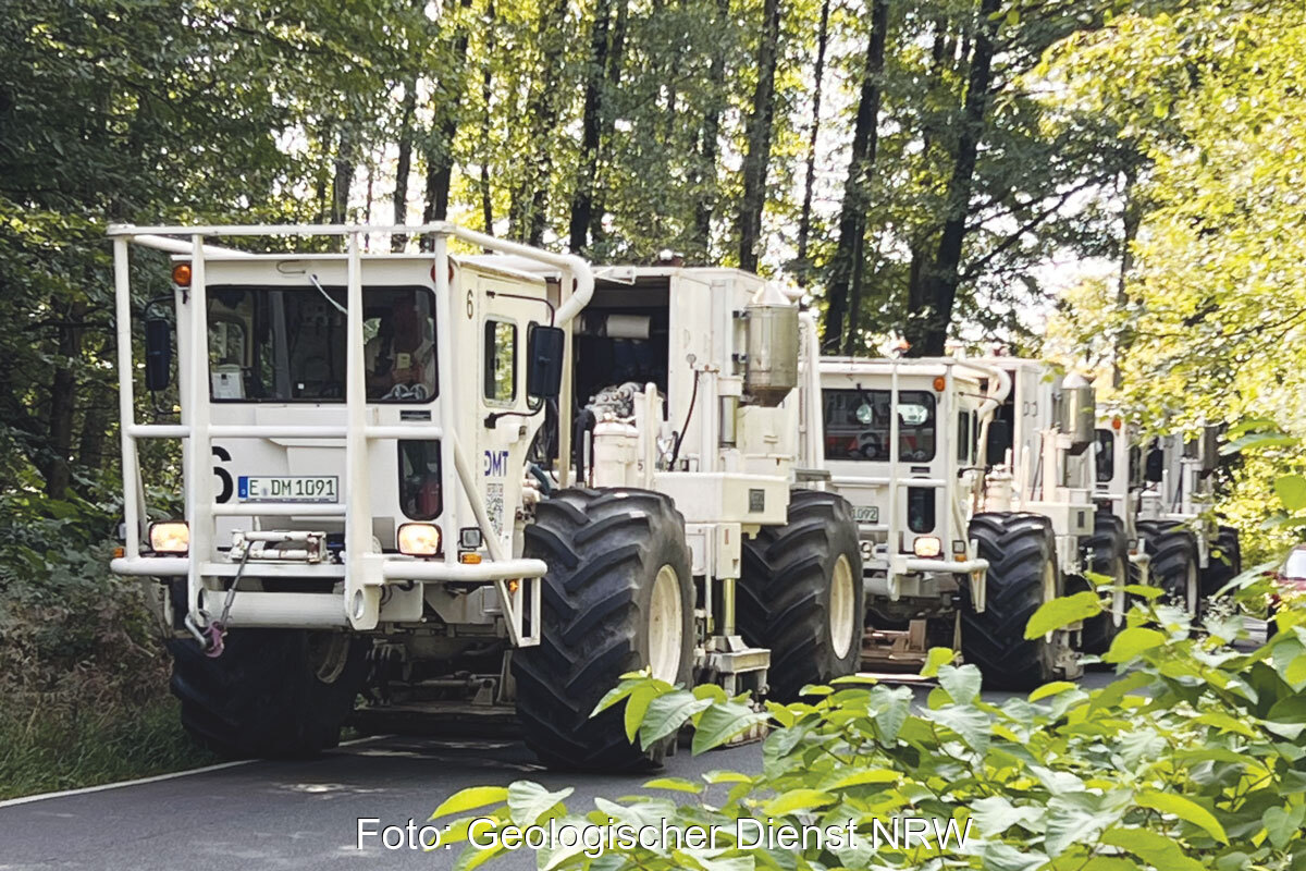 Weiß lackierte Kraftwagen mit großen Reifen in einer Reihe auf einem Waldweg. Vibro-Trucks erkunden den Untergrund, um das Potenzial für Tiefengeothermie-Bohrungen zu erkunden. Das Geothermie-Beschleunigungsgesetz macht das künftig einfacher.