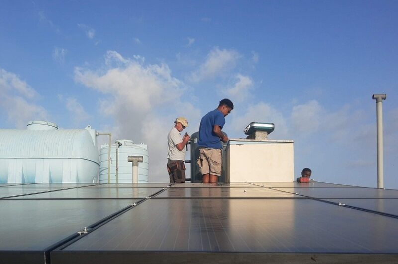 Mehrere Personen hinter einer Solardachanlage sowie Produktionstürmen im Hintergrund.