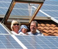 Eine Familie mit Baby blickt aus einem Dachfenster. Um sie herum ist das Dach mit Solarmodulen gedeckt.