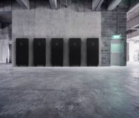 In einer leergeräumten Fabrikhalle stehen fünf große schwarze Batteriespeicher an einer Wand.