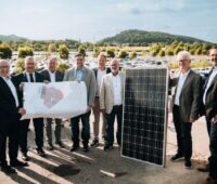 8 Männer mit Plan udn PV-Modul vor im Hintergrund stehenden Autos.