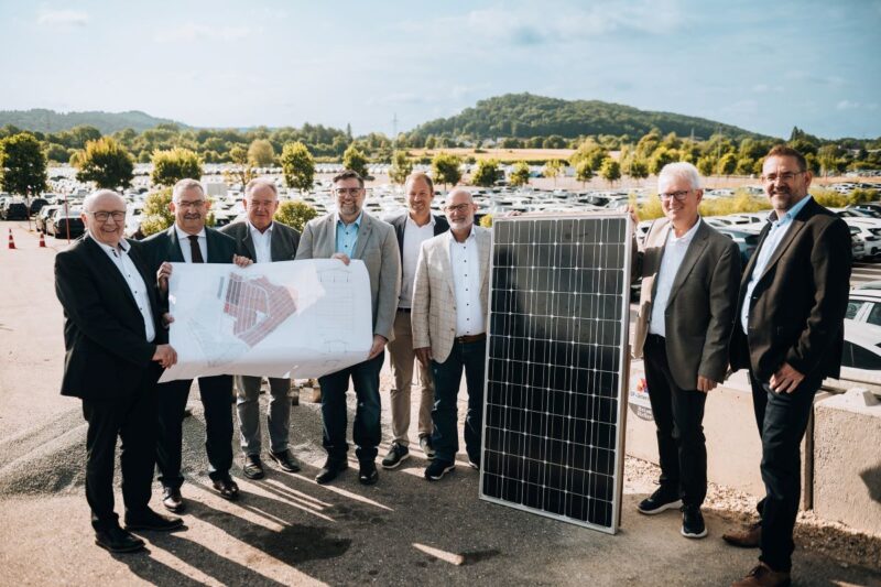 8 Männer mit Plan udn PV-Modul vor im Hintergrund stehenden Autos.