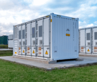 Verschiedene Batteriespeichersysteme in Containern stehen neben und hintereinander auf Fundamenten im Rasen.
