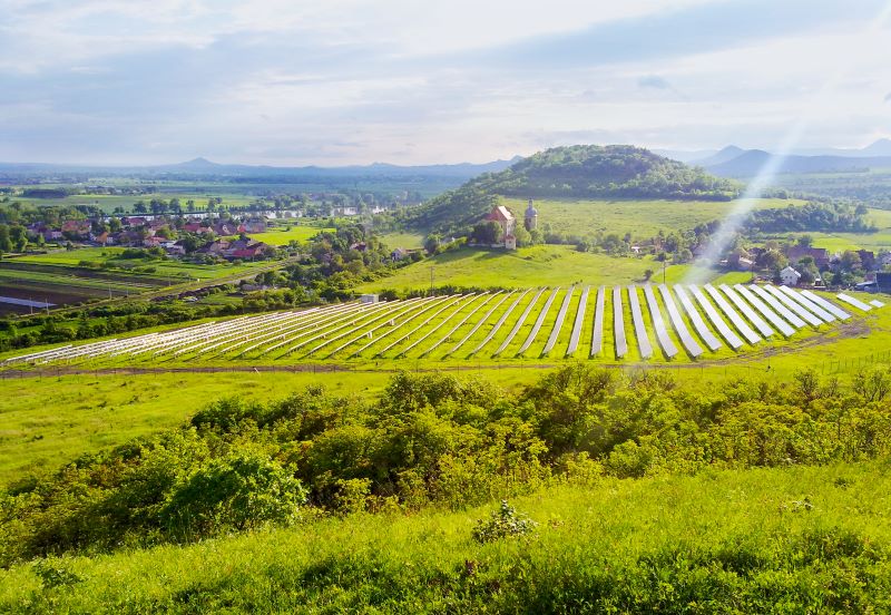 Auf einem Solarpark in Tschechien inmitten grüner Wiesen und mit einem alten Dorf und Kirche im Hintergrund strahlt die Sonne herab.